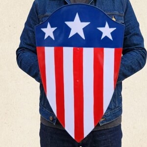 May include: A shield with a pointed bottom featuring a blue upper section with three white stars and a lower section with alternating vertical red and white stripes. The shield has a smooth, glossy finish and is held in front of a blue denim jacket.