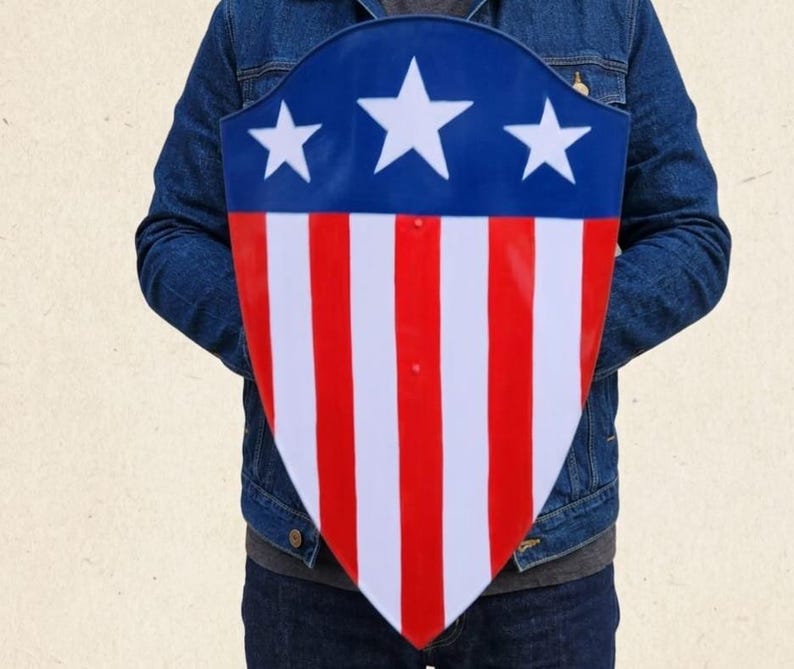 May include: A shield with a pointed bottom featuring a blue upper section with three white stars and a lower section with alternating vertical red and white stripes. The shield has a smooth, glossy finish and is held in front of a blue denim jacket.