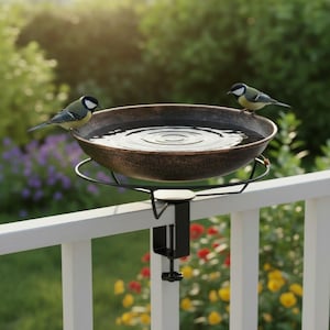 May include: A bronze-coloured bird bath with a black metal frame and clamp, attached to a white railing. Two small birds perch on the edge of the bath, which is filled with water. The background features green foliage and colourful flowers.