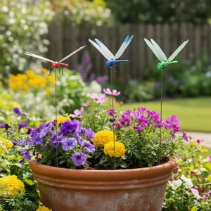 Puede incluir: Tres estacas de jardín con forma de libélula de colores rojo, azul y verde se exhiben en una maceta de terracota llena de vibrantes flores moradas, amarillas y rosas. Las libélulas tienen alas delicadas y están montadas en varillas de metal.