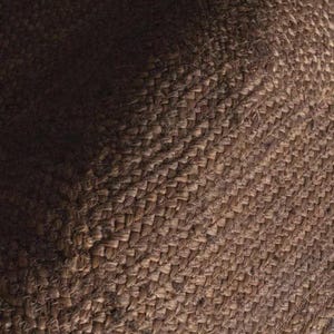 May include: A close-up view of a brown woven textile featuring a dense, interlocking basketweave pattern. The material displays a natural, fibrous texture with variations in light and shadow across the surface, highlighting the thick, sturdy weave.