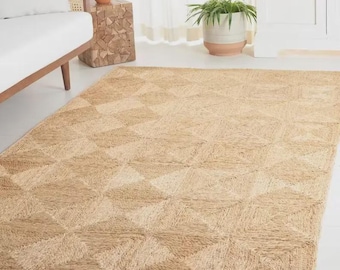 May include: A rectangular area rug crafted from natural tan jute fibers, featuring a repeating geometric pattern of squares and triangles woven into the surface. The rug has a textured, coarse appearance with a consistent light brown color throughout.