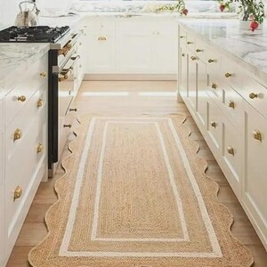 May include: A rectangular jute rug with a scalloped edge and a white border. The rug is placed in a kitchen with white cabinets and a marble countertop. The rug's dimensions are approximately 24 inches wide and 72 inches long.
