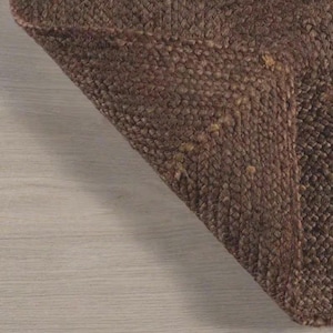 May include: A dark brown rug with a thick, braided, and textured weave. The material features a dense, interlocking pattern of fibers that create a raised, tactile surface. The corner of the rug is folded over, revealing the consistent weave.