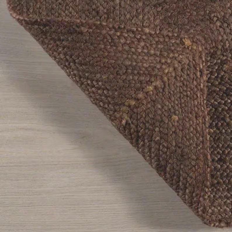 May include: A dark brown rug with a thick, braided, and textured weave. The material features a dense, interlocking pattern of fibers that create a raised, tactile surface. The corner of the rug is folded over, revealing the consistent weave.