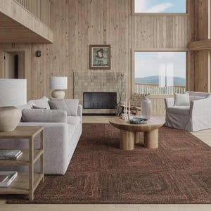 May include: A living room featuring a light gray sofa, a matching armchair, and a round wooden coffee table. A wooden side table with a lamp sits in the foreground. The room has light wood paneled walls, a fireplace, and a dark brown area rug.
