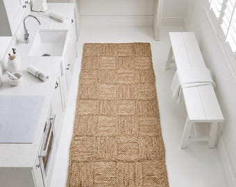 May include: A rectangular runner rug made of natural tan jute fibers, featuring a woven checkerboard pattern with alternating square sections of vertical and horizontal textures, positioned on a white floor in a kitchen setting.