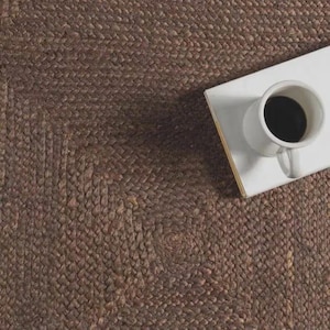 May include: A dark brown braided jute rug with a textured surface. A white rectangular coaster sits on the rug, supporting a white ceramic mug filled with black coffee. The mug features a handle on the right side and a circular opening at the top.