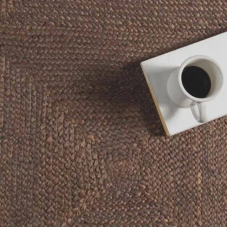 May include: A dark brown braided jute rug with a textured surface. A white rectangular coaster sits on the rug, supporting a white ceramic mug filled with black coffee. The mug features a handle on the right side and a circular opening at the top.