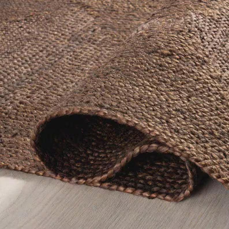 May include: A dark brown area rug featuring a thick, woven, natural fiber construction with a visible braided edge. The rug is folded over to reveal the dense, textured surface and the sturdy, interlaced pattern of the material throughout.