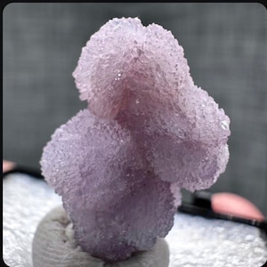 May include: A close-up of a lavender-colored mineral specimen. The crystalline structure has a textured, granular appearance. The mineral is mounted on a white surface, with a gray background.