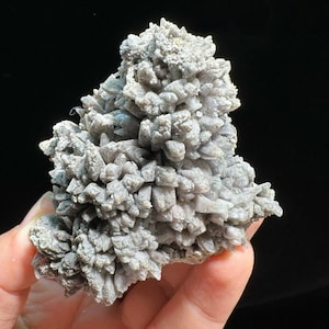 May include: A cluster of light gray crystals with a rough, textured surface. The crystals are densely packed, forming a layered, irregular shape. The background is black, highlighting the mineral's details.
