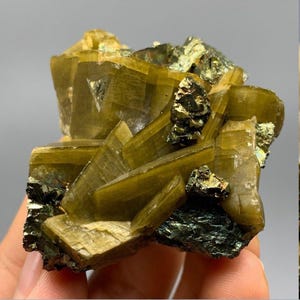 May include: A natural mineral specimen featuring a cluster of olive-green crystals with a layered, fan-like structure. The crystals are accented with small, dark, metallic-looking inclusions. The overall form is rough and irregular.