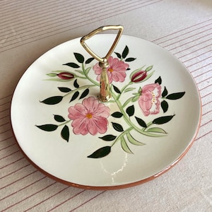 Puede incluir: Una bandeja de servicio de dos niveles con una superficie blanca, con diseños florales rosas y hojas verdes. La bandeja tiene un asa dorada y un borde marrón. Una pieza decorativa para servir alimentos o exhibir artículos.