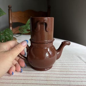 May include: A brown ceramic coffee dripper and pot set featuring a cylindrical top section with a handle and a rounded base with a spout. The glossy, solid brown glaze covers the entire exterior of the two-piece stackable brewing vessel.