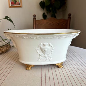Puede incluir: Un cuenco ovalado de cerámica blanca con borde dorado y cuatro patas de garra doradas. El exterior presenta un relieve blanco de una urna central flanqueada por pájaros y guirnaldas florales, con un patrón de enredadera en la parte superior.