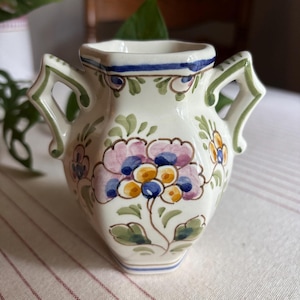 May include: A small cream-colored ceramic vase with two handles, featuring hand-painted floral motifs in shades of purple, yellow, and green, accented by blue horizontal bands around the rim and base, with a hexagonal body shape.