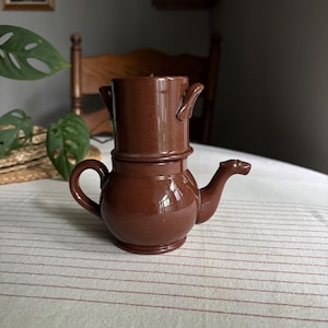 May include: A glossy brown ceramic coffee dripper set consisting of a rounded base pot with a side handle and a protruding spout, topped with a cylindrical filter holder featuring two small side handles. The entire piece has a uniform dark brown finish.