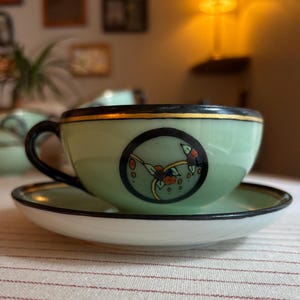 May include: A pale green ceramic teacup and saucer set featuring a black rim with a thin gold band. The cup displays a central circular black medallion containing a stylised floral motif in orange, blue, and green, complemented by a black handle.