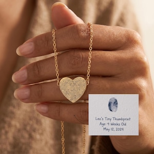 Op de afbeelding: Een gouden ketting met een hartvormige hanger met een vingerafdrukpatroon. De ketting wordt in een hand gehouden. Een kaart toont een vingerafdruk en de tekst: "Leo's Tiny Thumbprint, Age: 4 Weeks Old, May 12, 2024."