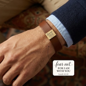 May include: A brown leather bracelet with a gold-coloured rectangular accent. The accent features the text "fear not FOR I AM WITH YOU Isaiah 41:10" in black script. The bracelet is worn on a wrist.