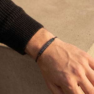 May include: A black bracelet with a rectangular bar engraved with the Roman numeral "MMXXIV". The bracelet is made of a dark metal and is worn on a wrist. The background is a neutral color.