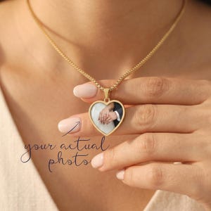 May include: A gold-toned heart-shaped pendant necklace featuring a custom photo insert of two hands holding each other. The pendant hangs from a delicate gold-toned chain. The text your actual photo is written in black script with an arrow pointing.