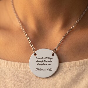 May include: A silver-toned circular pendant necklace featuring the engraved text I can do all things through him who strengthens me. [Philippians 4:13]. The pendant is suspended from a matching silver-toned metal chain with circular links.