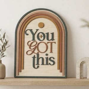 Pu&ograve; includere: Una targa da parete in legno a forma di arco con un design ad arcobaleno a strati nei toni del verde scuro, terracotta e giallo senape. La targa riporta la scritta "You GOT this" in rilievo con lettere verde scuro e color terracotta.