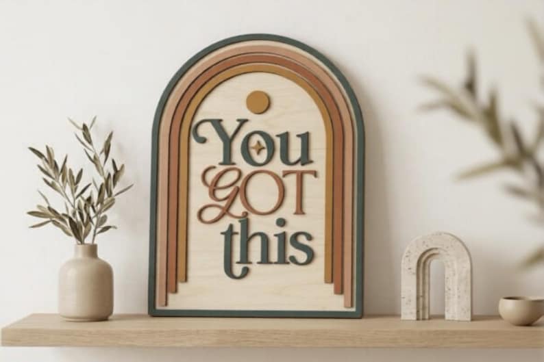 Pu&ograve; includere: Una targa da parete in legno a forma di arco con un design ad arcobaleno a strati nei toni del verde scuro, terracotta e giallo senape. La targa riporta la scritta "You GOT this" in rilievo con lettere verde scuro e color terracotta.