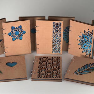 May include: A collection of wooden journals with intricate laser-cut designs. The journals feature various patterns, including a moon, flowers, and feathers, with blue accents. The covers are bound with metal rings, creating a unique and artistic aesthetic.