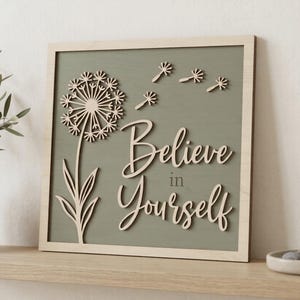 Pu&ograve; includere: Una targa da parete quadrata in legno con sfondo verde salvia chiaro e cornice in legno naturale. Presenta un disegno di tarassaco tagliato al laser con semi fluttuanti e il testo "Believe in Yourself" in carattere corsivo di legno in rilievo.