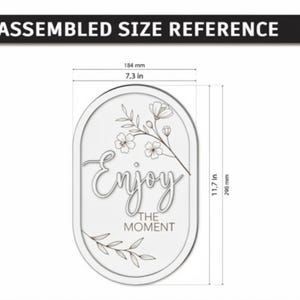 Pu&ograve; includere: Un cartello ovale con il testo "Enjoy THE MOMENT" e disegni floreali a linee. Le dimensioni sono 184 mm di larghezza per 298 mm di altezza. Il design include un ramo fiorito nella parte superiore e un ramoscello di foglie in quella inferiore.