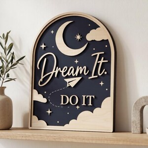 Pu&ograve; includere: Una targa da parete in legno ad arco con sfondo blu scuro, caratterizzata da intagli in legno chiaro a forma di luna crescente, nuvole, stelle, un aeroplanino di carta e il testo "Dream It. DO IT" in un mix di corsivo e stampatello.