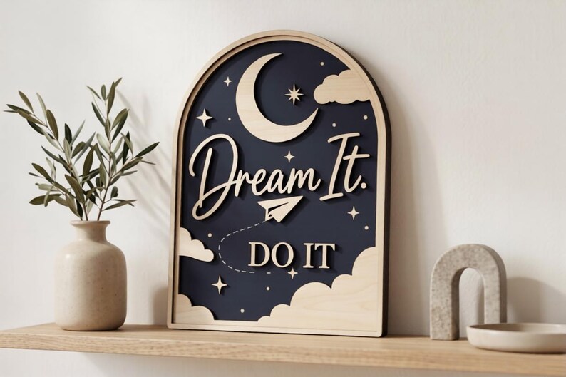 Pu&ograve; includere: Una targa da parete in legno ad arco con sfondo blu scuro, caratterizzata da intagli in legno chiaro a forma di luna crescente, nuvole, stelle, un aeroplanino di carta e il testo "Dream It. DO IT" in un mix di corsivo e stampatello.
