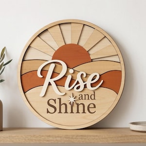 Pu&ograve; includere: Un cartello da parete circolare in legno con un design a strati raffigurante un sole e un paesaggio nei toni del legno chiaro, beige e terracotta. Il testo "Rise and Shine" &egrave; in caratteri corsivi e serif in rilievo con una stella.