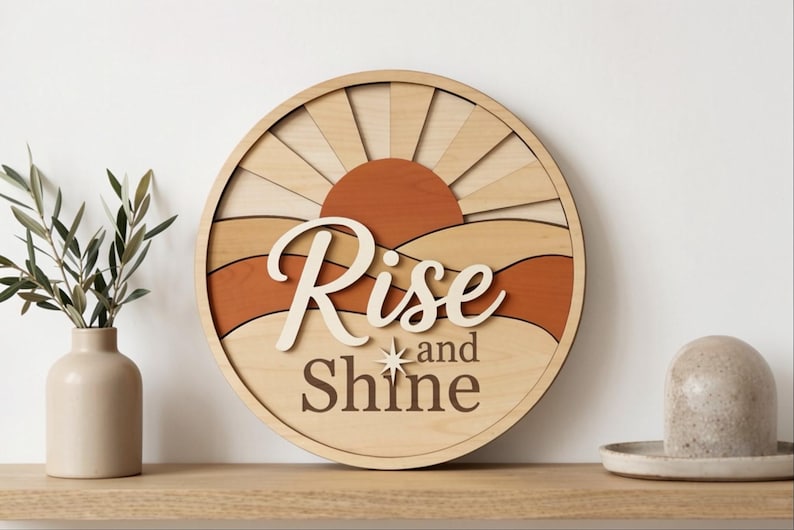 Pu&ograve; includere: Un cartello da parete circolare in legno con un design a strati raffigurante un sole e un paesaggio nei toni del legno chiaro, beige e terracotta. Il testo "Rise and Shine" &egrave; in caratteri corsivi e serif in rilievo con una stella.