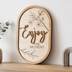 Pu&ograve; includere: Una targa da parete ovale in legno con la scritta Enjoy tagliata al laser e il testo THE MOMENT stampato. La targa in legno chiaro &egrave; decorata con delicate illustrazioni a linee di steli floreali e foglie posizionate sopra e sotto il testo.