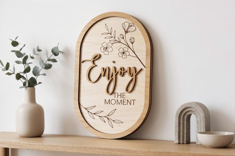 Pu&ograve; includere: Una targa da parete ovale in legno con la scritta Enjoy tagliata al laser e il testo THE MOMENT stampato. La targa in legno chiaro &egrave; decorata con delicate illustrazioni a linee di steli floreali e foglie posizionate sopra e sotto il testo.