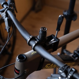 May include: Close-up of a bicycle handlebar with a black finish. The bike has a matte olive green frame and a black tool holder with a screwdriver and bits. The handlebar has the text "DTD" and "SANTA" printed on it.