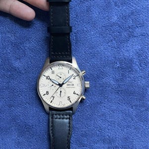 Pode incluir: Um relógio de pulso prateado com uma bracelete de couro preta. O mostrador do relógio é branco com algarismos e ponteiros pretos. O relógio tem o texto "IWC" e "SKR 20" impresso no mostrador. O relógio está sobre uma superfície azul texturizada.