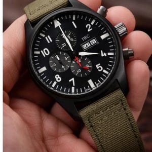 Op de afbeelding: Zwart polshorloge met een zwarte wijzerplaat, witte cijfers en drie sub-wijzerplaten. Het horloge heeft een groene canvas band. De wijzerplaat toont het merk "IWC" en de dag van de week.