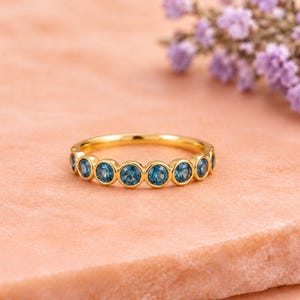 Può includere: Un anello a fascia in oro con una serie di pietre preziose blu rotonde incastonate a lunetta, disposte in fila lungo la metà superiore della fascia. L'anello presenta una finitura metallica lucida e una forma circolare costante.