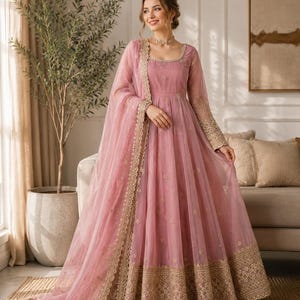 May include: A floor-length dusty pink gown featuring long sleeves, a square neckline, and a full skirt. The garment is adorned with intricate gold embroidery along the hem and cuffs, and includes a matching sheer dupatta with a scalloped gold border.
