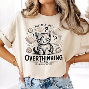 Puede incluir: Camiseta color crema con un gato de dibujos animados y el texto "MENTALLY BUSY" y "OVERTHINKING AGAIN IT'S A FULL-TIME JOB." El diseño incluye signos de interrogación y elementos de papel, creando un gráfico caprichoso.