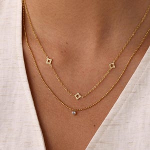 May include: A layered gold necklace set featuring two delicate chains. The top chain displays three small, square-shaped openwork charms, while the bottom chain features a single, tiny, round, light-colored stone pendant centered on a thin gold chain.