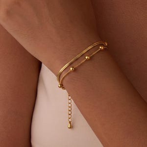 May include: A gold-toned double-strand bracelet featuring a flat snake chain and a delicate chain adorned with small, evenly spaced spherical beads. The bracelet includes an adjustable extender chain with a small teardrop-shaped charm at the end.