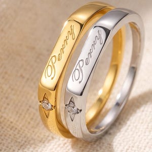 May include: Two rings, one gold-toned and one silver-toned, each featuring the engraved name Perry in a cursive script. Below the name, each ring displays a small, four-pointed star motif centered with a single, clear, round-cut gemstone.