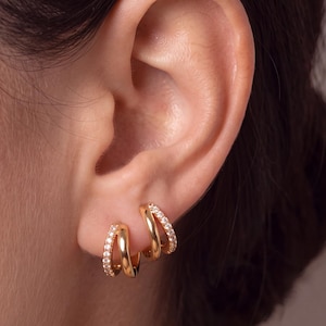 May include: A pair of gold hoop earrings featuring a double-band design. One band is smooth polished gold, while the other is encrusted with small, clear, sparkling stones. The earrings are worn in the earlobe, showcasing a layered, multi-hoop appearance.