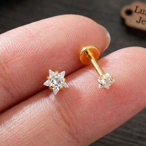 May include: A gold-tone flat back stud earring featuring a star-shaped clear gemstone setting. The jewellery piece has a thin post and a small circular base. The text LumiNova Jewelry is visible on a small brown tag in the background.